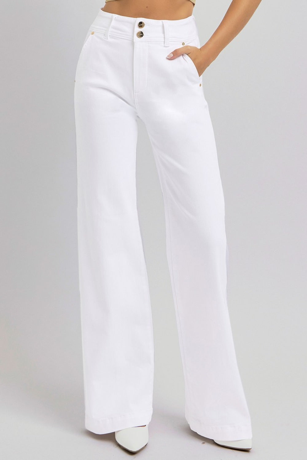 The lower body of a person is shown wearing RISEN Tummy Control Double Button Wide Leg Jeans, featuring a high-rise cut and wide-leg design in white. These jeans include both front pockets and tummy control, paired with pointed-toe white shoes against a plain light gray background.