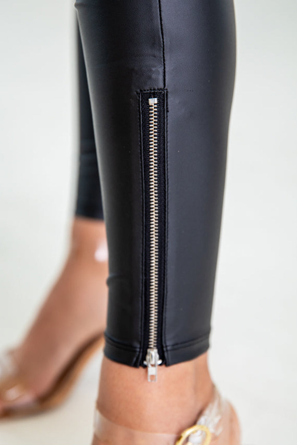 A close-up showcases a person wearing the Faux Leather Zipper Detail High Waist Leggings in black, featuring a striking silver zipper on the lower leg. The outfit is completed with clear, open-toe heels adorned with a gold buckle, all elegantly displayed on a light-colored surface.