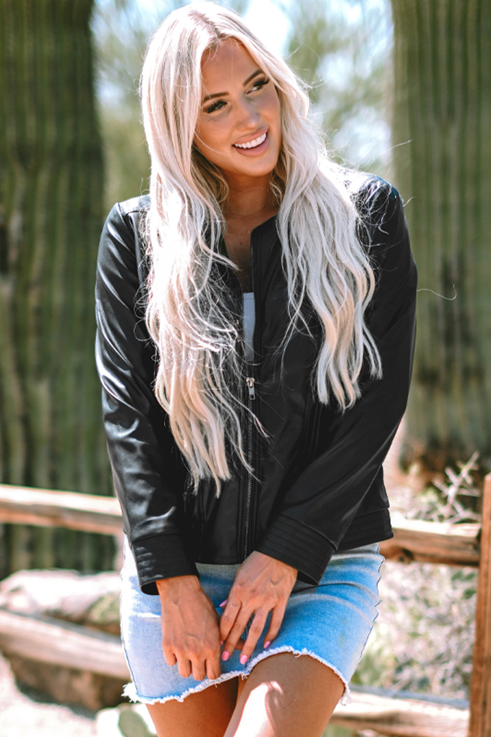 Young blond woman wearing faux leather black bomber jacket & denim skirt, front view, zipped